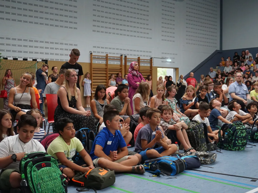 Kinder sitzen mit Ranzen auf dem Hallenboden.